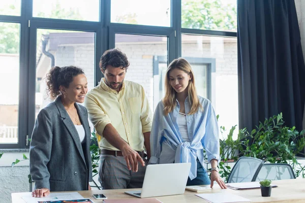 Businessman pointing at laptop near smiling interracial businesswomen in office — Stock Photo