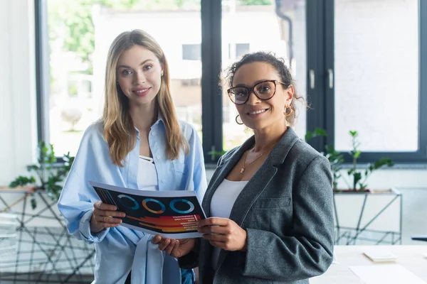 Happy multiethnic businesswomen holding paper and looking at camera in office — Stock Photo