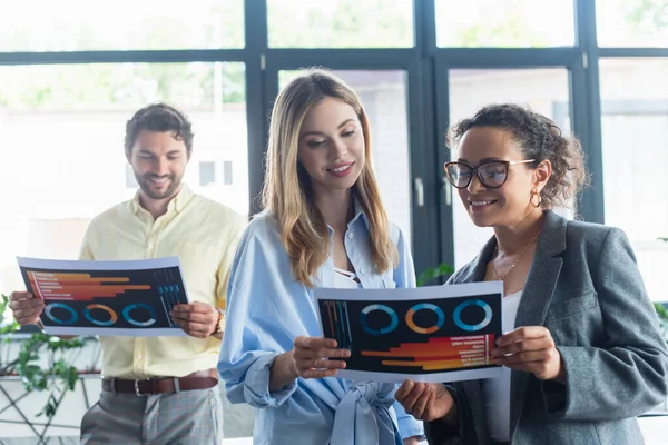 Smiling multiethnic businesswomen holding document near blurred colleague in office — Stock Photo