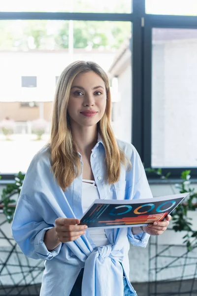 Jovem empresária sorrindo para a câmera enquanto segurava papel no escritório — Fotografia de Stock