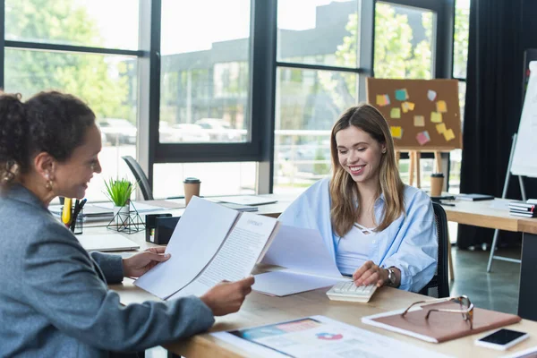 Cheerful businesswoman using calculator near blurred african american colleague in office — Stock Photo