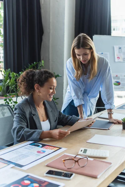 Smiling african american businesswoman holding paper folder near colleague in office — Stock Photo