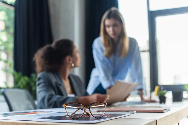 Eyeglasses on documents near blurred interracial businesswomen in office — Stock Photo