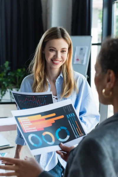 Smiling businesswoman holding document and looking at blurred african american colleague in office — Stock Photo