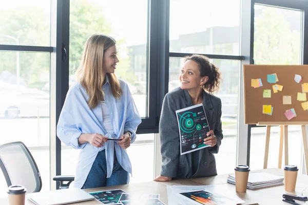 Positive multiethnic businesswomen working with documents near coffee to go in office — Stock Photo