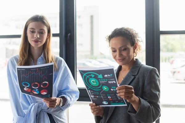 Smiling african american businesswoman looking at paper with charts near young colleague in office — Stock Photo