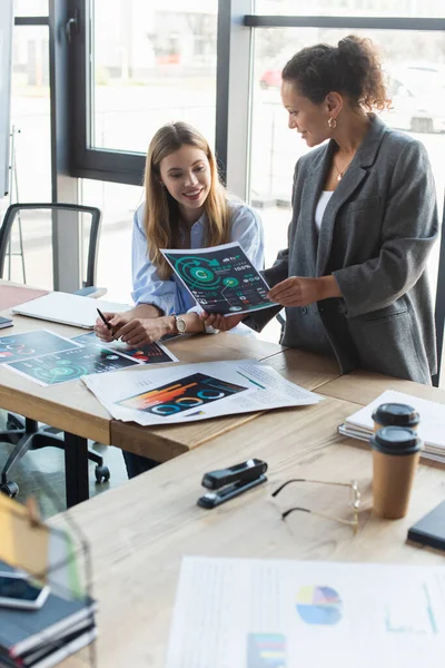 Lächelnde afrikanisch-amerikanische Geschäftsfrau hält Papier neben fröhlichen Kollegen in der Nähe von Coffee to go und Dokumenten im Büro — Stockfoto