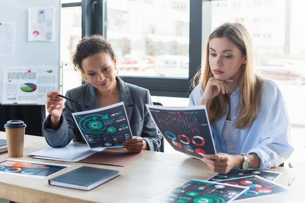 Multiethnic businesswomen working with documents near coffee and blurred flipchart in office — Stock Photo