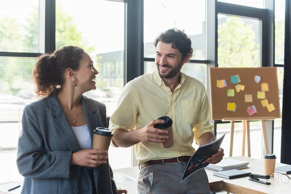 Cheerful interracial business people with coffee to go and document talking in office — Stock Photo