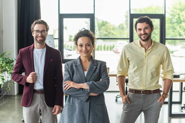 Sorrindo afro-americana empresária de pé com braços cruzados perto de colegas no escritório — Fotografia de Stock