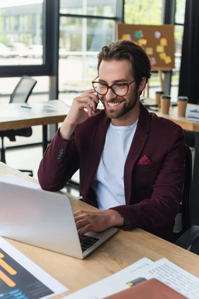 Jeune homme d'affaires en lunettes parlant sur smartphone et utilisant un ordinateur portable au bureau — Photo de stock