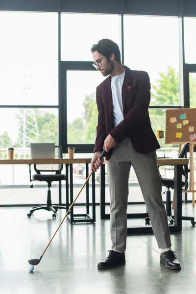 Vista lateral do jovem empresário em óculos jogando golfe no escritório — Fotografia de Stock