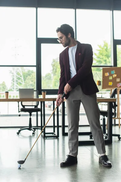 Vista lateral de un joven hombre de negocios en gafas jugando al golf en la oficina - foto de stock