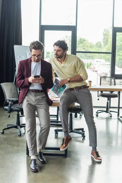 Businessmen with paper using smartphone near table in office — Stock Photo