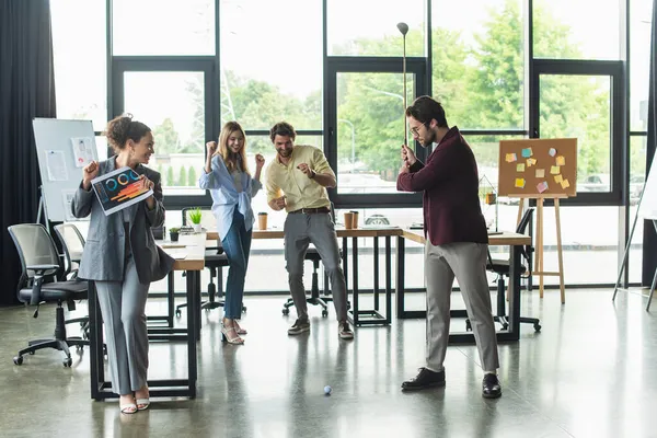 Businessman playing golf near excited multiethnic colleagues in office — Stock Photo