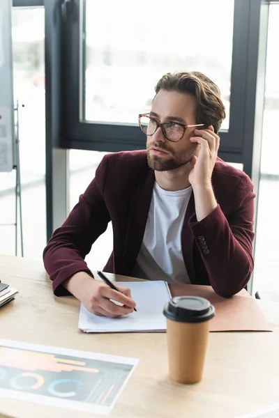 Pensive businessman writing on paper near blurred coffee to go in office — Stock Photo