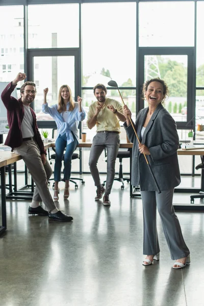 Excited african american businesswoman holding golf club near blurred excited colleagues in office — Stock Photo
