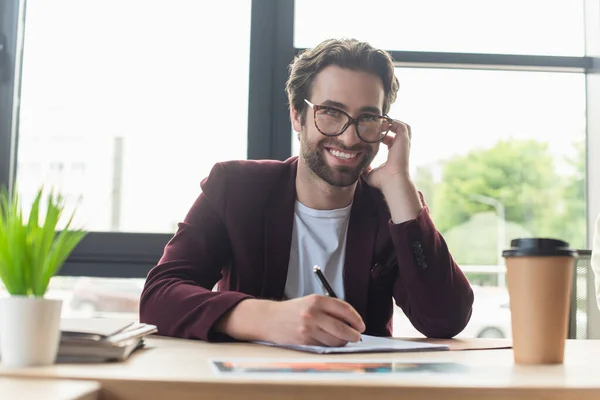 Positive businessman in eyeglasses writing on paper near blurred takeaway drink in office — Stock Photo