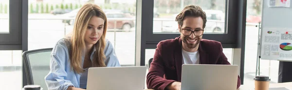 Hombre de negocios sonriente en gafas usando portátil cerca de colega en la oficina, pancarta - foto de stock