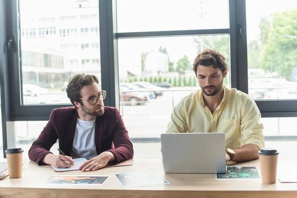 Uomini d'affari che scrivono su carta e utilizzano laptop vicino a caffè e documenti in ufficio — Foto stock