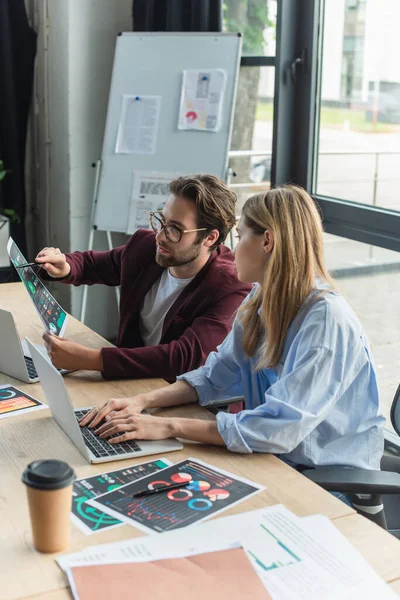 Empresario señalando el papel cerca de colega usando portátil en la oficina - foto de stock