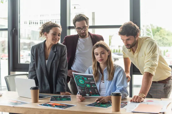 Geschäftsfrau hält Dokument mit Diagrammen neben Kollegen, Laptop und Kaffee im Büro — Stockfoto