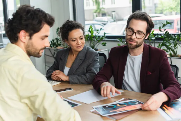 Jovem empresário apontando para documentos perto de colegas inter-raciais no escritório — Stock Photo