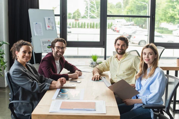 Gente de negocios multicultural positiva mirando la cámara cerca de papeles y calculadora en la oficina - foto de stock