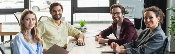 Des hommes d'affaires interraciaux joyeux regardant la caméra tout en travaillant au bureau, bannière — Photo de stock