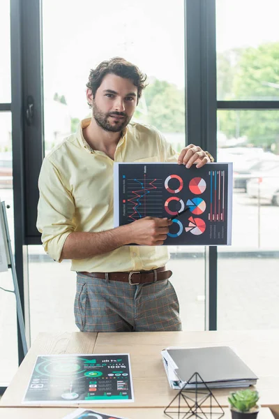 Businessman pointing at charts on paper and looking at camera in office — Stock Photo