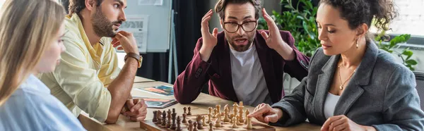 Astonished businessman sitting near interracial colleagues playing chess in office, banner — Stock Photo