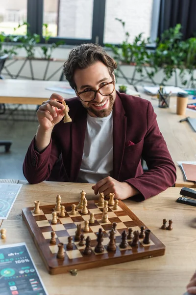 Homme d'affaires souriant dans des lunettes tenant une figure d'échecs près de l'échiquier dans le bureau — Photo de stock