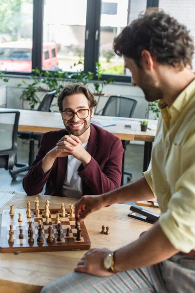 Smiling businessman in eyeglasses looking at blurred colleague playing chess in office — Stock Photo