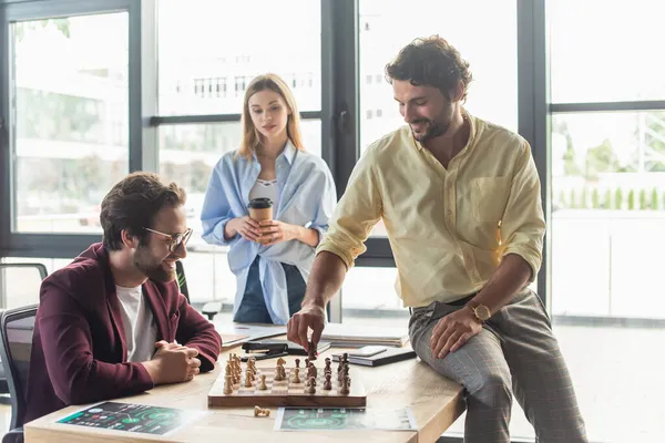 Lächelnde Geschäftsleute beim Schachspielen neben verschwommenem Kollegen mit Kaffee im Büro — Stockfoto