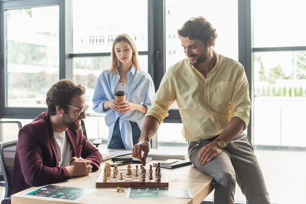 Des hommes d'affaires souriants jouent aux échecs près d'un collègue flou avec du café au bureau — Photo de stock