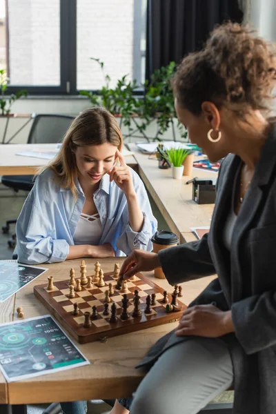 Pensive businesswoman looking at chess on board while playing with african american colleague in office — Stock Photo