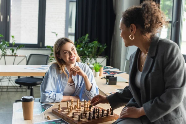 Femme d'affaires souriante regardant un collègue afro-américain jouer aux échecs près des papiers et du café au bureau — Photo de stock