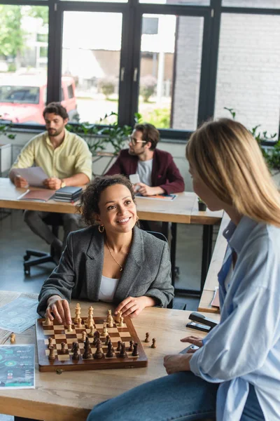 Міжрасові бізнесмени грають в шахи біля розмитих бізнесменів в офісі — стокове фото