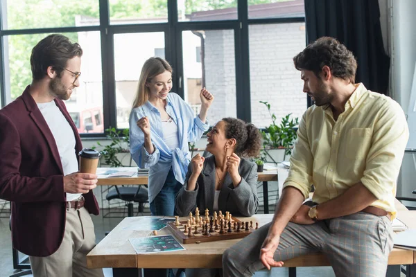 Interracial businesswomen showing yes gesture near colleagues playing chess in office — Stock Photo