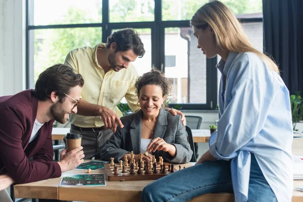 Улыбающаяся африканская американская бизнесвумен играет в шахматы рядом с коллегой, указывая пальцем и деловыми людьми в офисе — стоковое фото