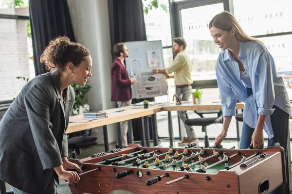 Alegre interracial businesswomen jugando futbol de mesa mientras borrosa colegas trabajando en oficina - foto de stock