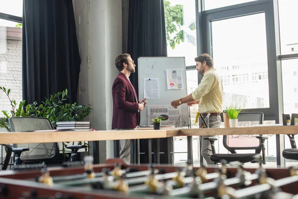 Side view of businessmen working with papers on flipchart near blurred table football in office — Stock Photo