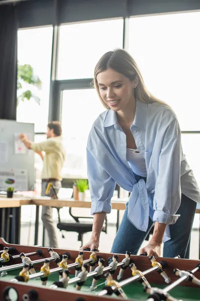 Mujer de negocios sonriente jugando al futbol de mesa cerca de colega borroso en la oficina - foto de stock