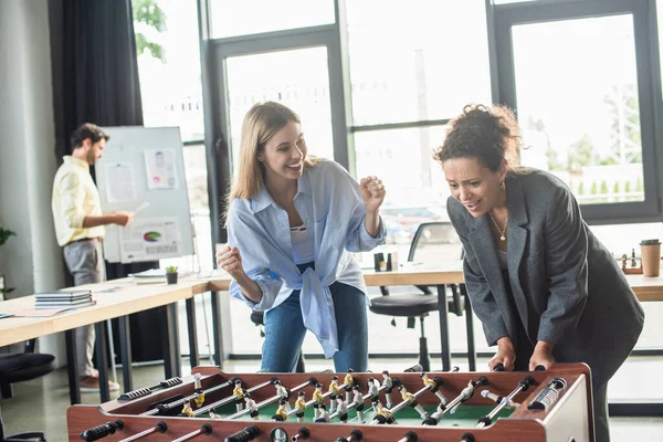 Excited businesswoman showing yes gesture near african american colleague playing table soccer in office — Stock Photo
