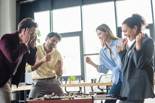 Gente de negocios multiétnicos emocionados jugando fútbol de mesa cerca triste colega en la oficina - foto de stock