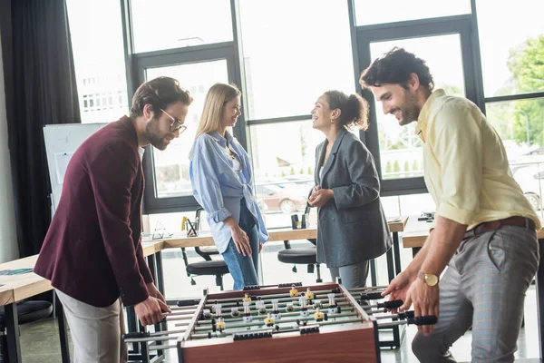 Businessmen playing table soccer near interracial colleagues talking in office — Stock Photo