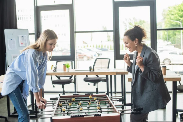 Empresária afro-americana entusiasmada mostrando sim gesto enquanto joga futebol de mesa com colega no escritório — Fotografia de Stock