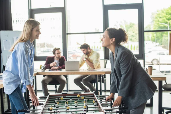 Fröhliche Geschäftsfrauen beim Tischkicker neben verschwommenen Geschäftsleuten im Büro — Stockfoto