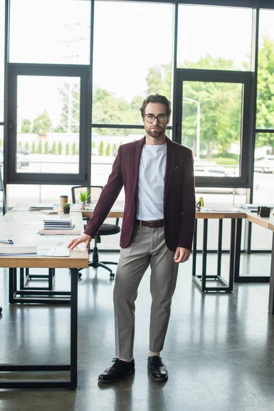 Jeune homme d'affaires en tenue de cérémonie debout près de la table de travail au bureau — Photo de stock