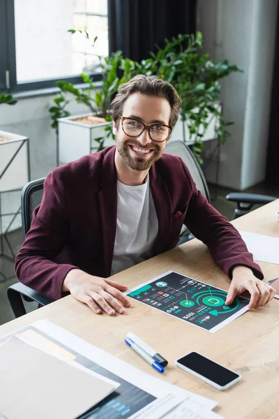 Happy businessman looking at camera near smartphone and papers on table in office — Stock Photo